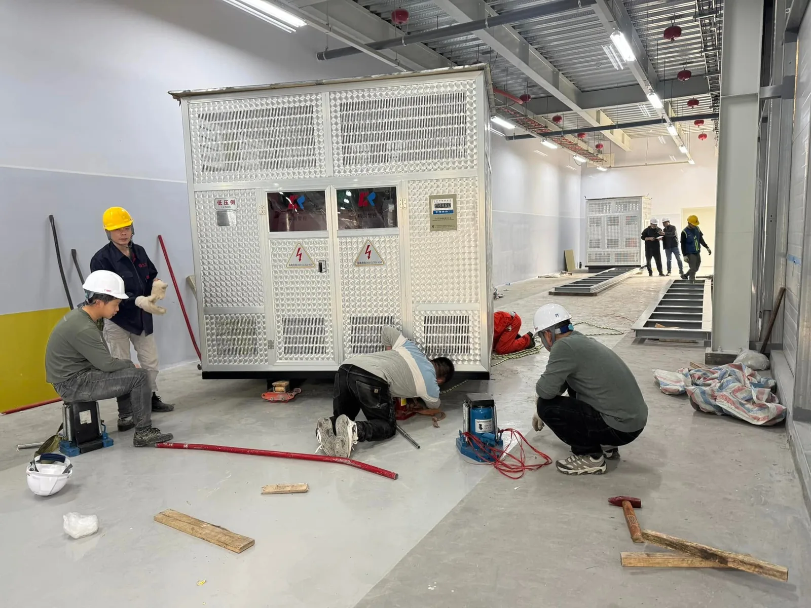 Thợ dùng kích thủy lực và con lăn căn chỉnh máy biến áp khô vào bệ trong phòng điện tại Xe Nâng Đức Anh