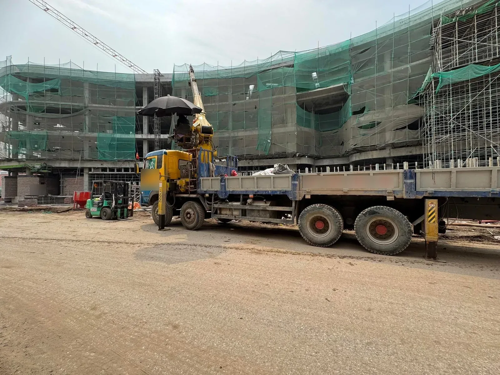 Xe tải cẩu chở thép đỗ trước công trình mặt cong có giàn giáo lưới xanh tại Xe Nâng Đức Anh